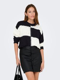 Pulover tricotat în navy și alb, cu un model geometric îndrăzneț; asortat cu o fustă din piele neagră, cu nasturi pe față. Poartă o geantă neagră în mână.