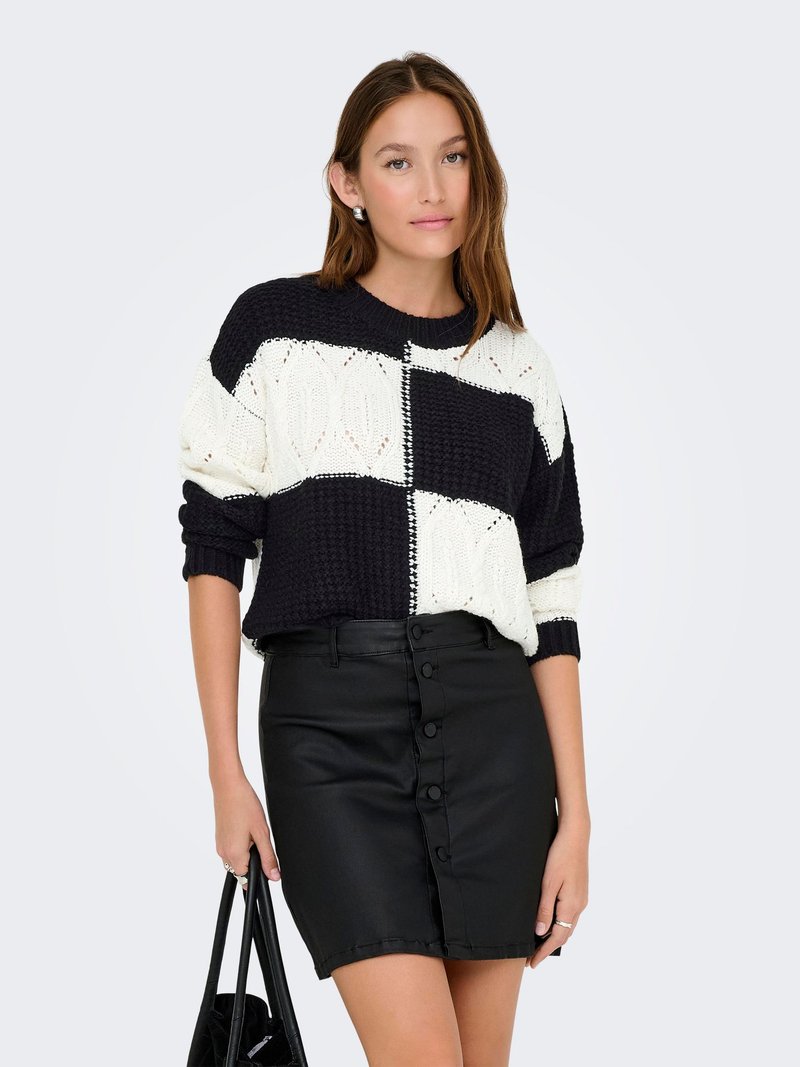 Pulover tricotat în navy și alb, cu un model geometric îndrăzneț; asortat cu o fustă din piele neagră, cu nasturi pe față. Poartă o geantă neagră în mână.