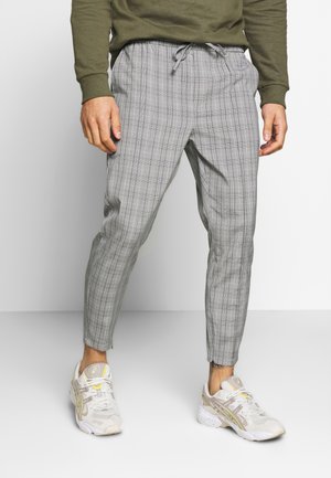 Mann, der graue Karo-Jogginghosen mit Kordelzug und weiße Turnschuhe trägt, steht vor einem schlichten weißen und grauen Hintergrund.