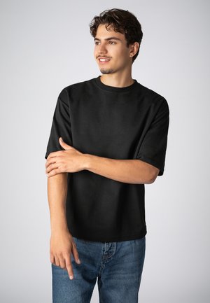 Schwarzes T-Shirt mit kurzen Ärmeln aus weicher Baumwolle. Mit rundem Ausschnitt und lockerer Passform. Kombiniert mit blauen Jeans.