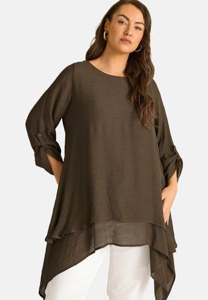 Femme portant une tunique asymétrique ample, brun foncé, avec des manches trois-quarts retroussées sur un pantalon blanc, debout les mains dans les poches.