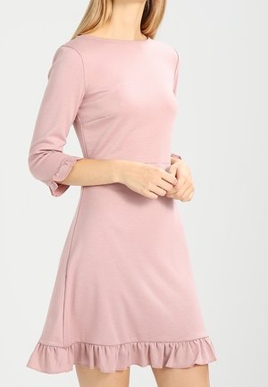 Kobieta w jasno różowej sukience do kolan z rękawami 3/4 i falbankami na rękawach i dole.
