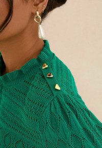 Pull en maille verte avec un motif texturé, comportant trois boutons en forme de cœur dorés à l'épaule et un col côtelé.