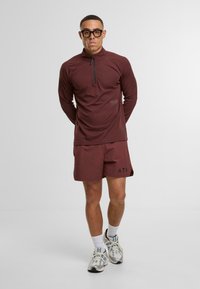 Bordeauxrotes sportliches Langarm-Oberteil mit Halb-Reißverschluss und passenden Shorts; strukturierten Stoff; schwarze Logo-Akzente; getragen mit weißen Socken und Laufschuhen.