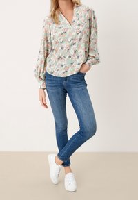 Blusa floreale in colori pastello con scollatura a V, maniche lunghe a sbuffo e vestibilità morbida, abbinata a jeans blu aderenti e sneaker bianche.
