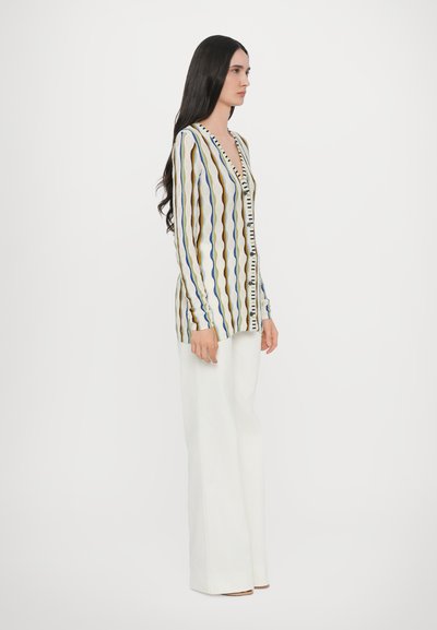 Stickad cardigan med vertikala vågiga ränder i blått, brunt och vitt. Har en V-ringning och knappfront. Styling med vita vida byxor.