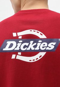 Camiseta de algodón roja con un gran logotipo en blanco y azul marino de "Dickies" que incluye un gráfico de herradura. Cuello redondo, mangas cortas, textura suave.