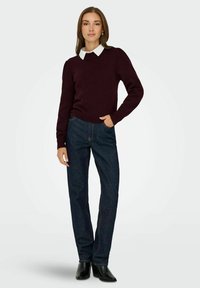 Maglione bordeaux con maniche a sbuffo, abbinato a jeans dritti blu scuro. Il modello indossa stivali neri con tacco e una camicia bianca con colletto.