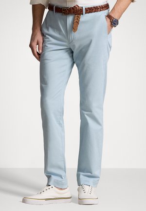 Mann mit hellblauer Hose, weißen schnürsenkellosen Sneakers, braunem geflochtenem Gürtel und Armbanduhr, eine Hand in der Hosentasche.