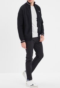 Marineblaue Zip-Jacke aus strukturiertem Stoff, mit hohem Kragen und kontrastierenden Streifenakzenten an den Bündchen. Kombiniert mit schwarzen Hosen und weißen Sneakers.