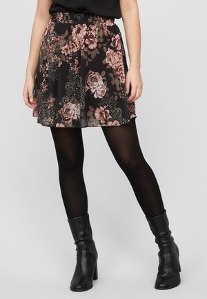 Kvinne iført et svart skjørt med blomster i rosa og brunt, svarte strømpebukser og svarte ankelstøvletter med hæl, på en ensfarget bakgrunn.