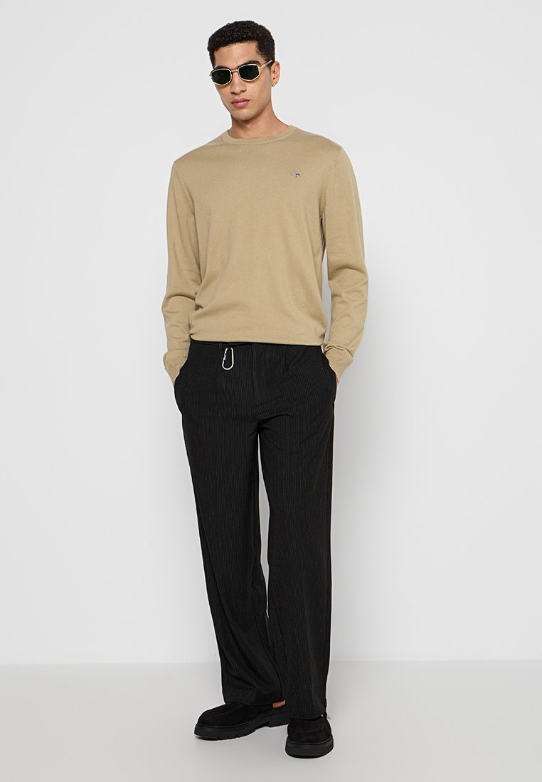 Beige Langarmhemd mit kleinem Logo, kombiniert mit schwarzen, gestreiften Hosen und schwarzen Slippern. Dunkle Sonnenbrillen runden das Outfit ab.