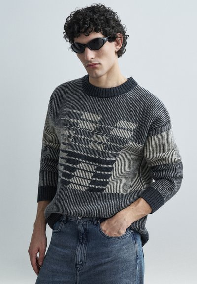 Pull en maille avec des motifs contrastants gris foncé et crème, col rond et poignets côtelés, assorti à un jean bleu.
