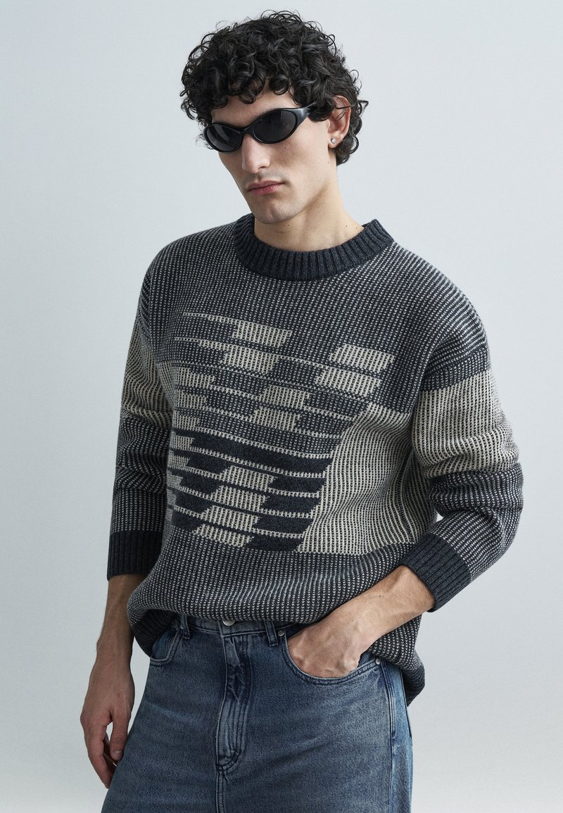 Camisola de malha com padrões contrastantes em cinzento escuro e creme, gola redonda e punhos canelados, combinada com jeans azuis.