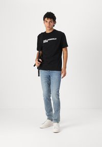Juoda medvilnės marškinėliai su balta "KARL LAGERFELD JEANS" logotipu, derinami su šviesiai mėlynomis džinsinėmis kelnėmis ir baltomis sportinėmis bateliais. Paprastas dizainas, trumpomis rankovėmis.