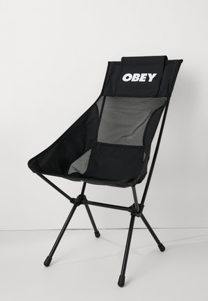 Scaun pliant negru cu spătar din țesătură tip plasă, logo-ul "OBEY" în partea de sus și un design triunghiular. Dispune de picioare din metal negru cu vârfuri din cauciuc.