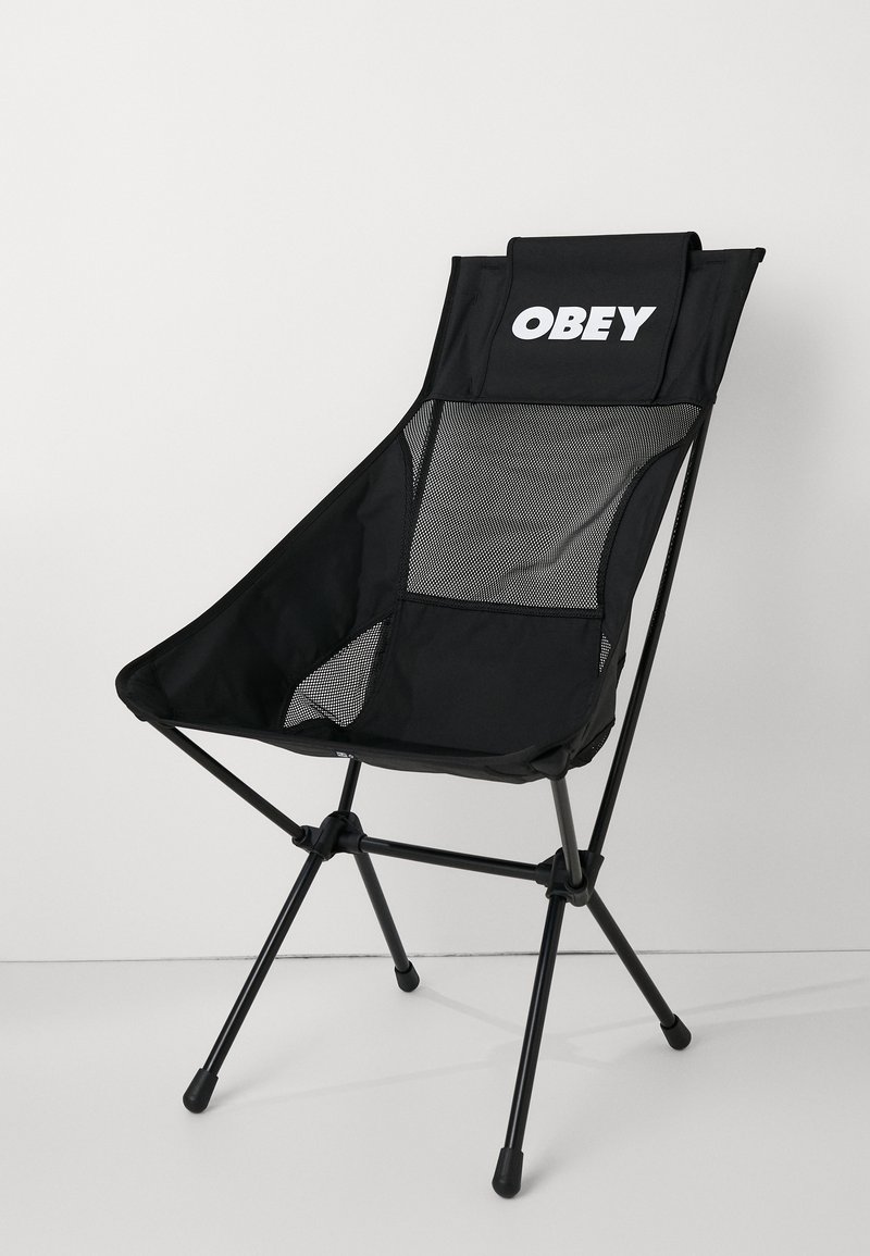 Črn zložljiv stol z mrežastim naslonjalom, na vrhu z logotipom "OBEY" in trikotno zasnovo. Ima črne kovinske noge z gumijastimi konicami.