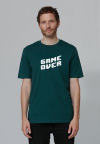 Donkergroen katoenen T-shirt met de tekst "GAME OVER" in vette, witte, blokletters; korte mouwen en een ronde hals.