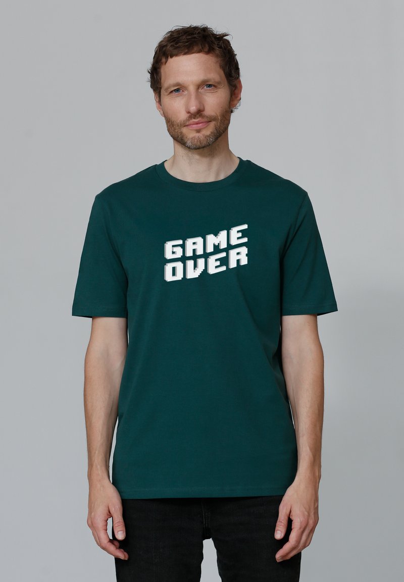 Donkergroen katoenen T-shirt met de tekst "GAME OVER" in vette, witte, blokletters; korte mouwen en een ronde hals.