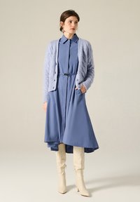 Abito azzurro chiaro a maniche lunghe con chiusura a bottoni e vita cintata, abbinato a un cardigan lavorato a maglia con texture di colore corrispondente e stivali beige al ginocchio.