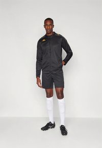 Fekete sportos melegítő cipzáras dzsekivel és rövidnadrággal, arany részletekkel. Fehér térd fölé érő zoknival és fekete stoplis cipővel viselve.