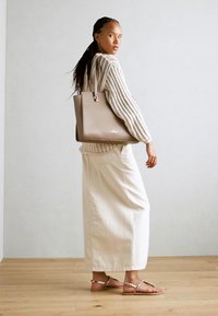 Beige leather tote with handles, ribbed cream sweater, cream long skirt, and rose gold sandals; wooden floor and neutral wall background.