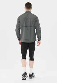 Grå lättvikts löparjacka med rak skärning, elastiska manschetter och en logotyp på ryggen, kombinerad med svarta korta leggings och sneakers.