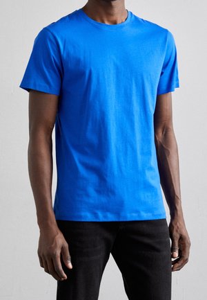 Homme portant un t-shirt uni bleu vif à manches courtes et un pantalon noir, debout devant un fond gris clair.