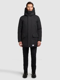 Muž stojící oblečený v černé zimní parce s kapucí a předními kapsami, černé džíny a černé boty na jednobarevném šedém pozadí.