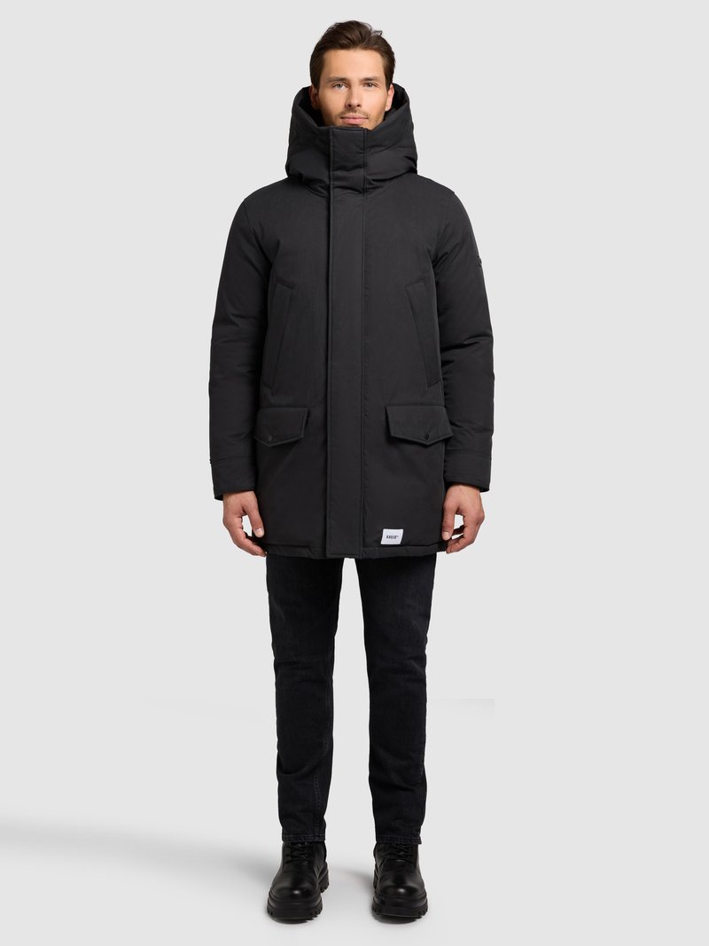 Muž stojící oblečený v černé zimní parce s kapucí a předními kapsami, černé džíny a černé boty na jednobarevném šedém pozadí.