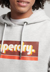 Sudadera gris con cordones, que presenta un bold logotipo en naranja y marrón. Textura suave, diseño casual con un ajuste relajado y un estilo en capas.