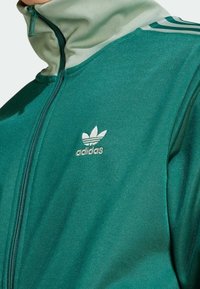 Grön zip-up jacka med hög krage, tillverkad av texturerat tyg. Har ljusgröna detaljer och broderad vit Adidas-logotyp.