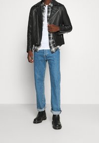 Giacca da motociclista in pelle nera sopra una camicia bianca, flanella a quadri, jeans azzurri chiari e stivali neri alla caviglia con orli grigi.