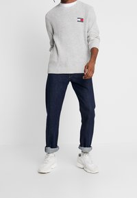 Grauer gerippter Pullover mit einem Logo-Patch, kombiniert mit dunkelblauen Jeans und weißen Sneakers. Auf den Jeans sind die aufgerollten Säume sichtbar.