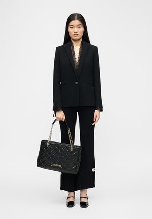 Kvinne iført en svart skreddersydd blazer og vide bukser, som holder en svart quiltet Love Moschino veske med gullkjedestropp.
