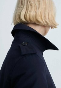Personne blonde aux cheveux courts portant un manteau bleu marine foncé avec un col montant et des détails de boutons, tournée vers la droite sur un fond uni.