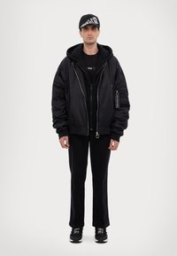 Giacca bomber nera sopra un hoodie nero, abbinata a pantaloni neri e sneaker. Un cappellino nero presenta testo bianco. Tessuto liscio e vestibilità comoda.