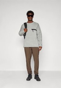 Sweatshirt cinzento texturizado com um bolso com fecho preto, combinado com calças castanhas e ténis pretos; o modelo carrega uma mochila preta.