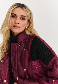 Bordový prešívaný pufér bez rukávov s čiernymi textúrovanými rukávmi a striebornými cvokmi, s kvetinovou podšívkou a vysokým golierom.