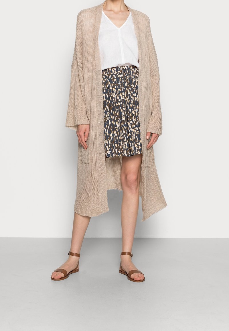 Femme portant un long cardigan en tricot beige par-dessus un chemisier blanc à col en V, une jupe plissée à motifs et des sandales marron à bride cheville, debout contre un mur blanc.