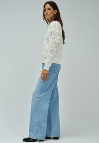 Sudadera blanca con detalle de encaje floral en las mangas, puños acanalados, combinada con pantalones vaqueros de pierna ancha color azul claro y botas de tobillo negras.
