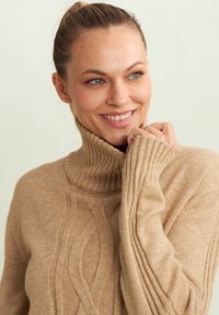 Pull en col roulé beige avec une texture à côtes, comportant un motif de vagues texturé à l'avant. Tissu doux, conception légère et manches ajustées.