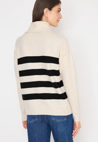 Beige Pullover mit hohem Kragen, ausgestattet mit auffälligen schwarzen horizontalen Streifen auf der Rückseite. Weiche Textur mit geripptem Saum und Bündchen.