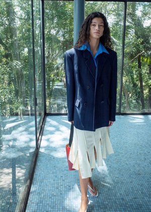 Vrouw met krullend haar die een oversized navyblauwe pinstripe blazer draagt, een blauw overhemd met kraag, een witte rok met een split, sandalen, en een rode handtas vasthoudt in een glazen kamer.