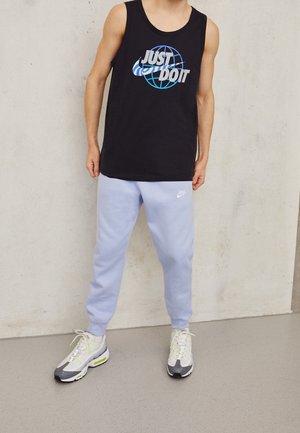 Personne portant un débardeur noir avec le texte "Just Do It" et un pantalon de survêtement Nike bleu clair, debout contre un mur clair et uni.