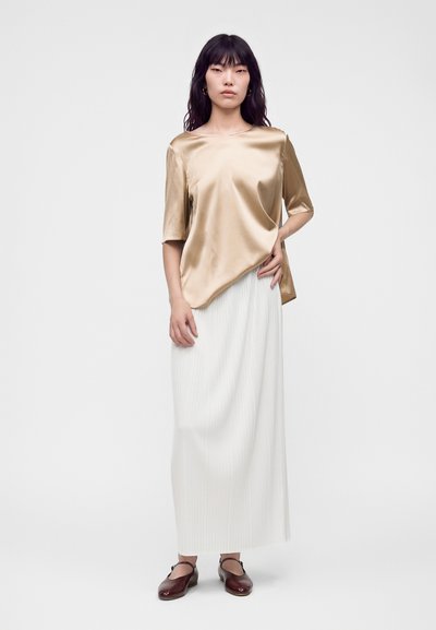 Femme aux longs cheveux foncés portant un haut beige brillant à manches courtes, une longue jupe plissée blanche et des chaussures plates marron foncé, debout devant un fond uni.
