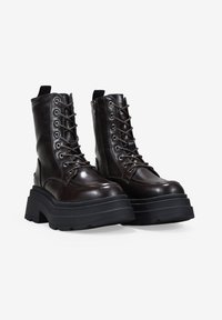 Par de botas de combate de cuero marrón oscuro con suelas de plataforma negras gruesas y cordones delanteros, mostradas sobre un fondo blanco.