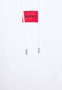Sudadera blanca con un parche rectangular rojo que presenta la palabra "HUGO" en negro. Cuerdas delgadas blancas con puntas de metal. Textura de tejido suave.