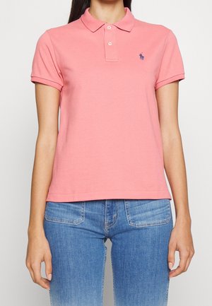 Kvinde iført en lyserød kortærmet poloshirt og blå jeans, står med armene afslappet ned langs siderne.