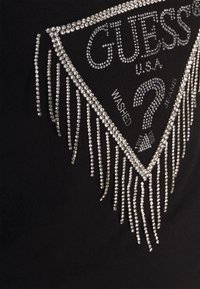 Musta t-paita, jossa on kolmiomainen strassikuvio ja hapsureunus, jossa lukee "GUESS USA", "WASHED" ja "JEANS".